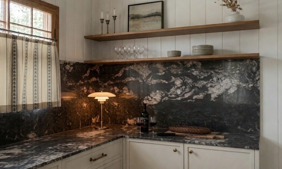 Kitchen corner with a dark veined stone splashback, oak shelves, a brass table lamp, cafe curtains and a lit candlestick
