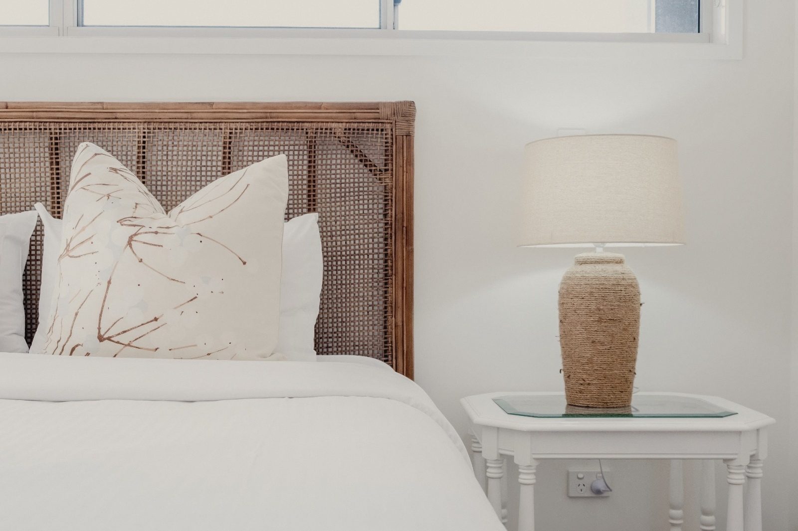 Not Nothing Beach House: bedroom with a woven rattan bedhead, white linen, a botanical cushion and a rope lamp on a white bedside table
