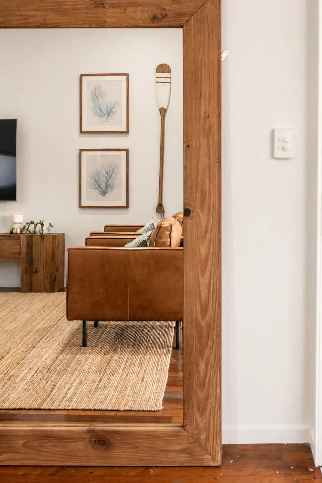 Not Nothing Beach House: timber-framed mirror reflecting a coastal living room with a leather armchair and jute rug
