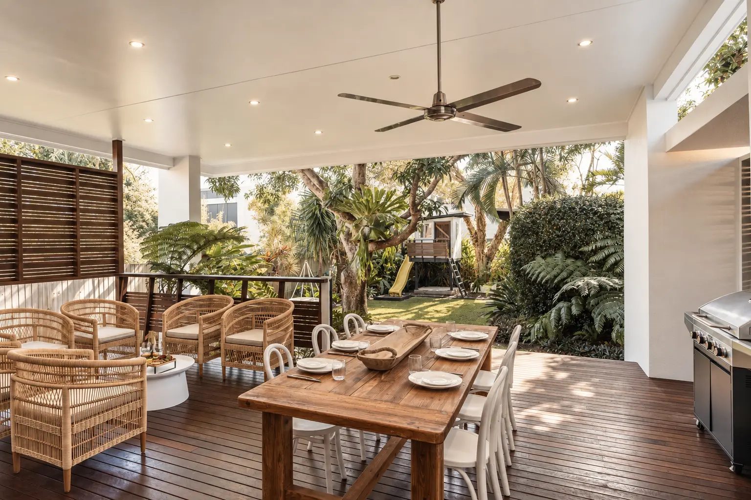 Covered timber deck with a long dining table, rattan lounge chairs and a leafy tropical garden beyond